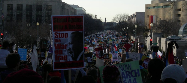 Eine Gruppe von Menschen, die auf einer Straße protestieren und Plakate halten, mit Gebäuden, Bäumen und Laternen im Hintergrund bei klarem Himmel.