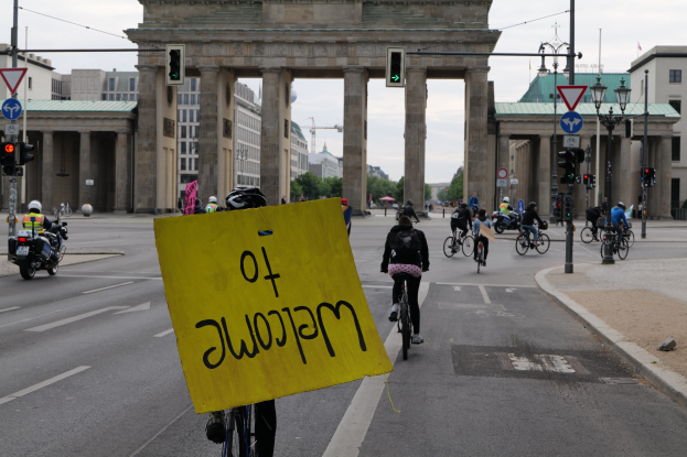 Gruppe von Menschen, die Fahrräder auf einer Straße vor dem Brandenburger Tor in Berlin, Deutschland, fahren, wobei eine Person ein gelbes Schild hält.