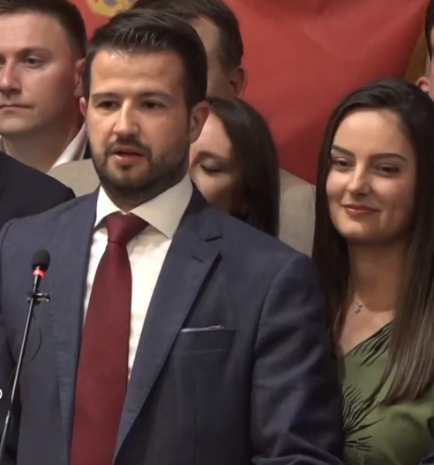 Ein Mann im Anzug und Krawatte steht an einem Rednerpult und hält eine Rede vor einer Menge mit einem Banner im Hintergrund, wahrscheinlich reagiert er auf ein Regierungsverbot von sozialen Medien.