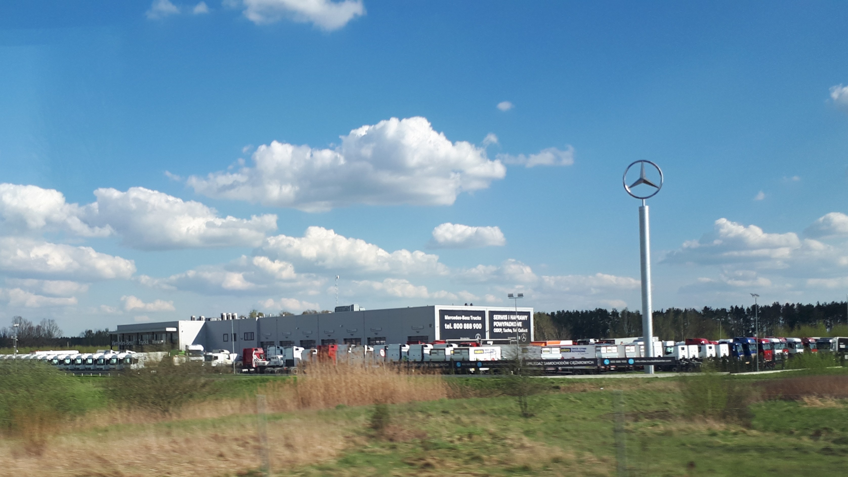 Mercedes-Benz-Fabrik in Deutschland mit Fahrzeugen auf der Straße, umgeben von Gras, Pflanzen, Bäumen, Gebäuden und Strommasten, einschließlich eines mit einem Logo.