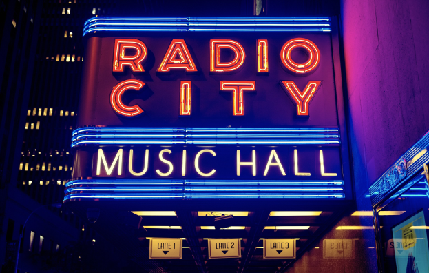 Das erleuchtete Radio City Music Hall in New York City mit hohen Gebäuden im Hintergrund, das eine 'Radio City'-Leuchtschrift und zusätzliche Schilder auf der rechten Seite zeigt.