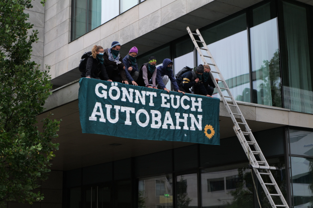 Gruppe von Menschen auf einem Gebäudedach mit einer Fahne, einem Baum links und Glasfenstern im Hintergrund, die scheinbar protestieren und die deutsche Regierung unterstützen.