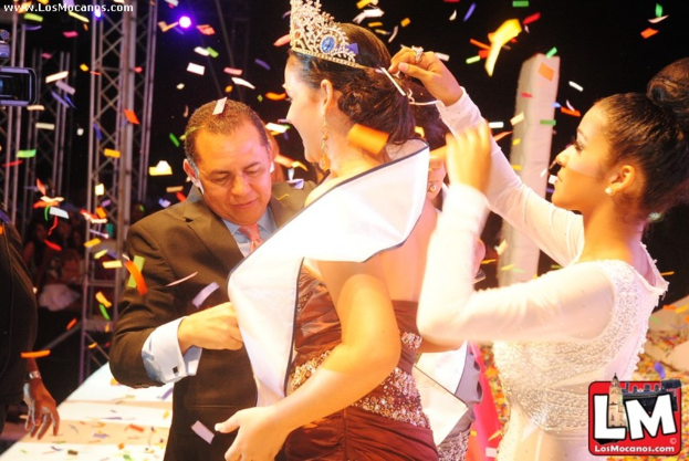 Ein Mädchen auf der Bühne bei einer Miss World Show, das eine Krone trägt, während ein anderer Mädchen sie von hinten anpasst und ein Mann in einem schwarzen Anzug vor ihr die Hand schüttelt, vor einem dunklen Hintergrund mit Metallrahmen.