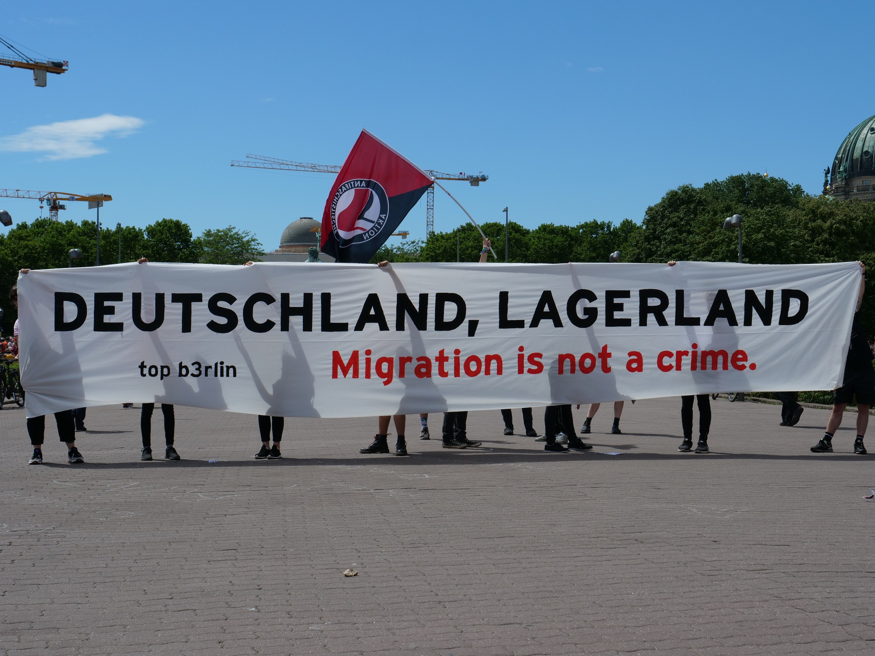 Menschen mit einem Banner, auf dem "Deutschland, Lagerland Migration ist kein Verbrechen" und einer Flagge, mit Straßeninfrastruktur und Gebäuden im Hintergrund.