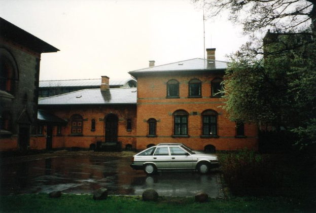 Ein Auto vor dem Auschwitz-Vernichtungslager, ein großes Backsteingebäude mit Fenstern und Türen, umgeben von Gras und Steinen, mit Bäumen auf der rechten Seite und einem klaren Himmel im Hintergrund.