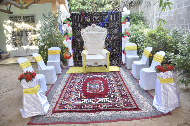 Hochzeitszeremonie im Garten mit weißen Stühlen, rotem Teppich, Blumenbouquets, Dekorationsgegenständen, Ballons, Pflanzen, Bäumen, einem Gebäude mit Fenstern und einer Tür sowie einer Wand auf der rechten Seite.