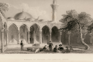 Eine Moschee in Payass mit Menschen, die auf dem Boden sitzen, umgeben von Bäumen, mit Text am unteren Bildrand.