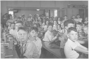 Schwarzes und weißes Foto von Kindern an langen Cafétiertafeln mit Tellern, Gläsern, Flaschen und anderen Gegenständen; eine Wand mit Fenstern und ein Schrank mit Gegenständen darauf sind im Hintergrund zu sehen.