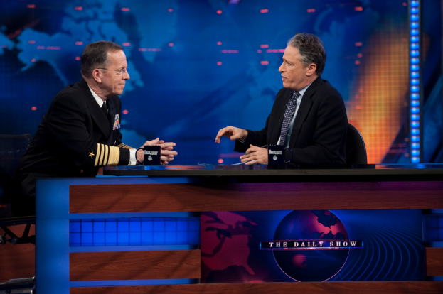 Zwei Männer in schwarzen Blazern, Hemden und Krawatten sitzen an einem Tisch mit Mikrofonen zwischen ihnen vor einem Fernseher und einem blauen Hintergrund.