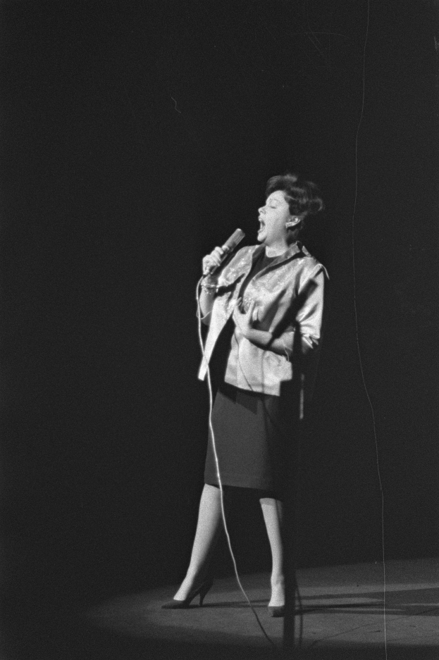 Eine Frau in einem schwarzen und weißen Outfit aus den 1950er-Jahren singt fröhlich auf einer von Scheinwerfern beleuchteten Bühne und hält ein Mikrofon in der Hand.