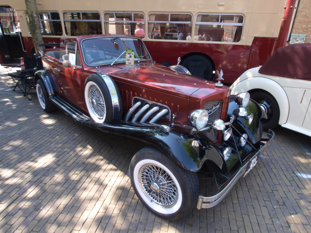 Ein Oldtimer neben einem Doppeldeckerbus auf einer Kopfsteinpflasterstraße geparkt, mit Stühlen und einem Baum im Vordergrund, einem Gebäude im Hintergrund und Menschen im Bus zu sehen.