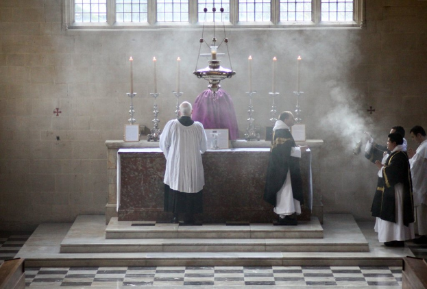 Eine Statue in der Mitte mit Kerzen auf Haltern, ein Sockel unten mit Menschen, eine Wand und Fenster im Hintergrund und ein Boden unten.