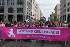 Eine Gruppe von Menschen marschiert auf einer Straße in Berlin mit einer pinken 'Happy Pride March'-Fahne, umgeben von Gebäuden, Laternenmasten und Verkehrszeichen unter einem bewölkten Himmel.