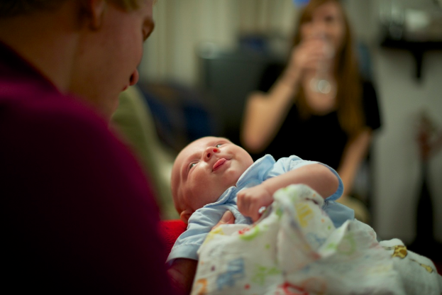 Ein Mann hält ein Baby in den Händen, vor ihm steht eine Frau und der Hintergrund ist unscharf.