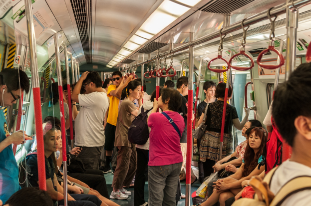 Menschen auf einem offenen Waggon eines Zuges sitzend oder stehend, mit Metallstangen, Griffen und Deckenleuchten.