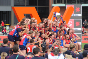 Eine Gruppe von Frauenfußballerinnen in blauen und roten Uniformen feiert mit einem Pokal vor einem jubelnden Publikum auf einem Rasenplatz.