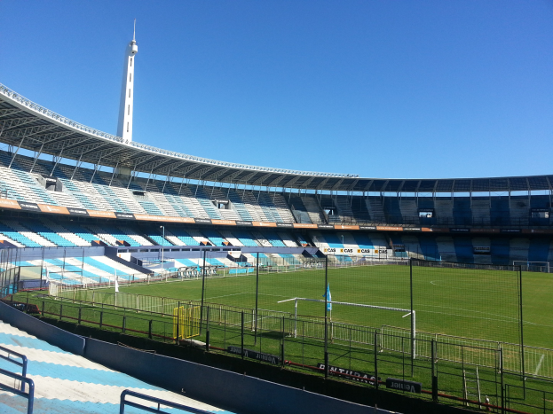 Großes Stadion mit einem Fußballfeld, umgeben von einem Zaun, einem Turm im Hintergrund und einem klaren blauen Himmel.
