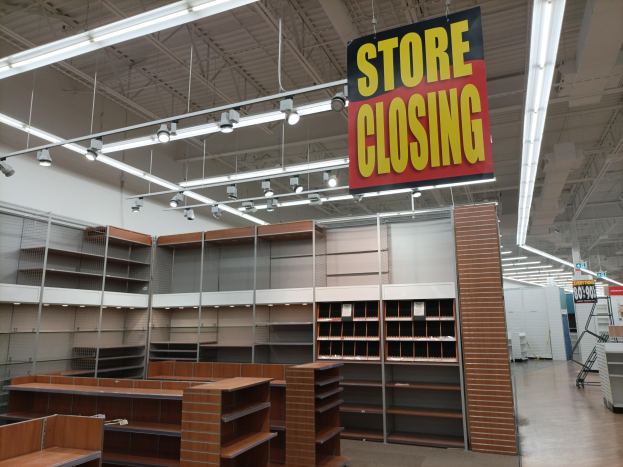 Store interior with a "Store Closing" sign hanging from the ceiling, shelves stocked with items, a text board, and overhead lighting.