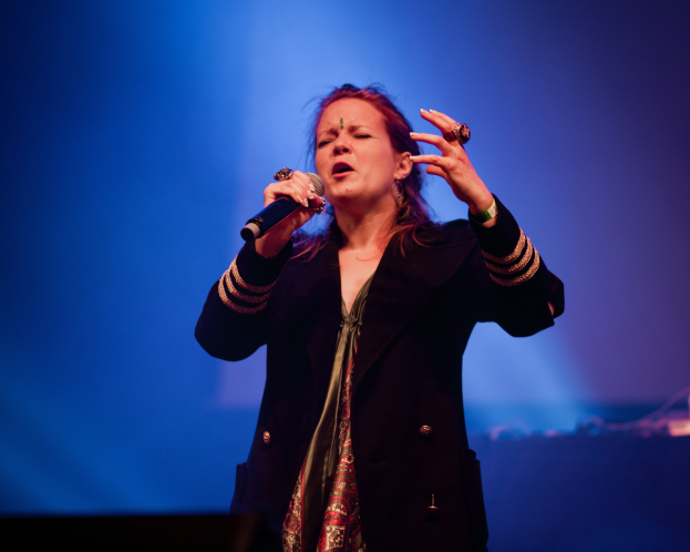 Eine Frau in einem schwarzen Kleid singt auf der Bühne in ein Mikrofon, beleuchtet von blauem Licht, ihre Augen geschlossen und ihr Gesichtsausdruck voller Emotion.