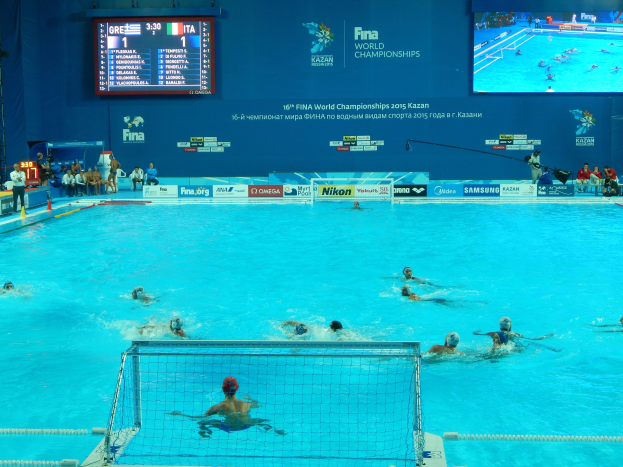 Wasserballspieler bei den FINA-Weltmeisterschaften 2016 in Kazan im Pool gegeneinander antretend, mit Zuschauern auf der linken Seite und einem Werbe-Scoreboard im Hintergrund.