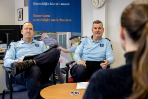 Zwei Polizeibeamte sitzen an einem Tisch mit einer Frau, umgeben von Papieren und Gegenständen, mit einem Wandbanner, einer Uhr und Fotorahmen im Hintergrund und einem weiteren Tisch mit einem Monitor und Zubehör links daneben.