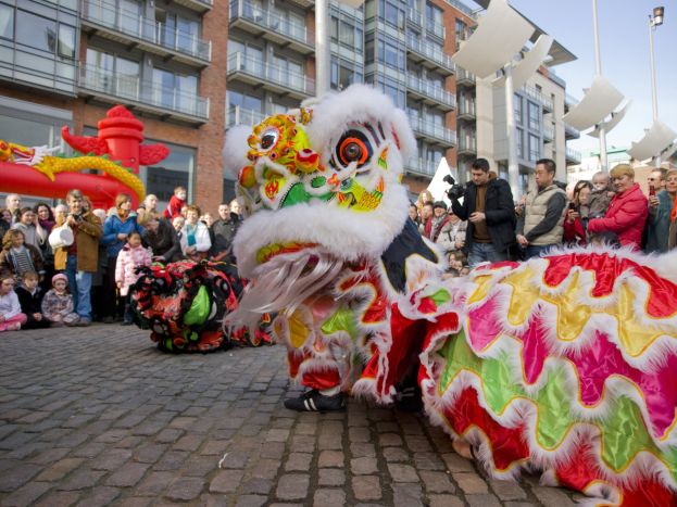 Ein lebhaftes chinesisches Neujahrsfest in Amsterdam mit einem Drachen tanzen vor einer Zuschauermenge, einige machen Fotos, vor einer Kulisse aus Gebäuden, Laternenmästen und einem klaren blauen Himmel.