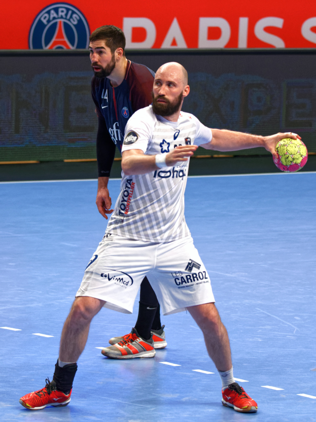 Zwei Männer spielen Handball auf einem Platz, einer hält einen Ball, mit einem Schild im Hintergrund, auf dem 'Paris Saint-Germain vs Paris Saint Germain' steht.