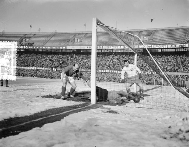 Ein Schwarz-Weiß-Foto eines Fußballspiels in einem Stadion, mit Spielern auf dem Boden, einer Person auf dem Schnee, einem Torpfosten, einer Fahne mit Text, Pfosten, Flaggen und einem bewölkten Himmel.