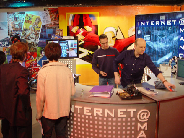 Mehrere Personen in einem Raum mit einem Tisch, auf dem sich eine Datei, eine Tastatur, ein Monitor und eine Maus befinden, sowie eine Wand im Hintergrund mit Plakaten und einer Cartoon-Figur auf dem Bildschirm.