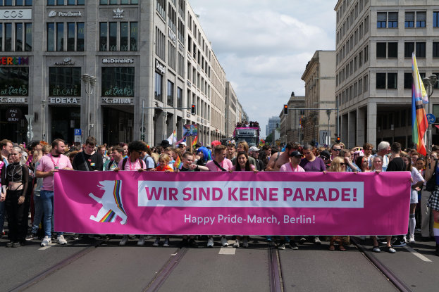 Eine Gruppe von Menschen marschiert auf einer Straße in Berlin mit einem pinken Banner mit der Aufschrift "Happy Pride March", während Gebäude, Laternenmasten und Verkehrszeichen die Straße säumen und der Himmel bewölkt ist.