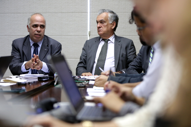 Ein Mann in einem blauen Hemd und Blazer steht in einer Ecke eines Konferenzraums und spricht mit vier sitzenden Männern, mit einem Tisch voller Gegenstände davor und einer weißen Wand dahinter.