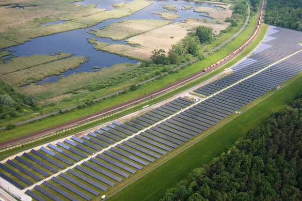 Luftaufnahme einer Solarpark mit Solarpanelen, umgeben von Bäumen, Gras, Pflanzen und Wasser, mit einem Zug auf einer nahen Eisenbahnschiene.