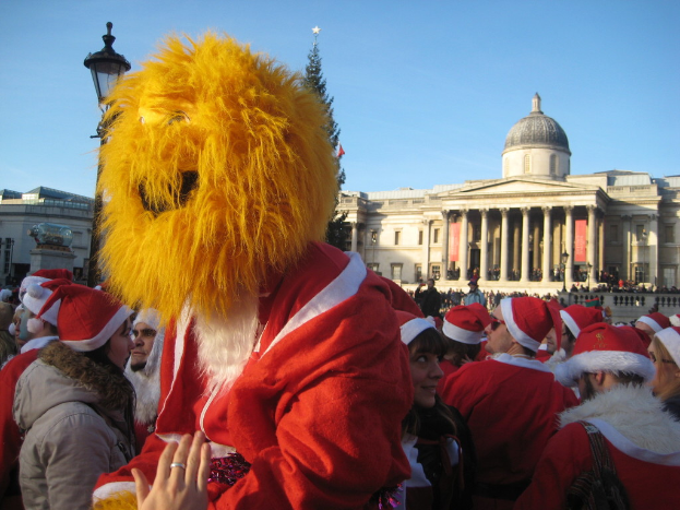 Person in Santa-Kostüm mit Tigermaske steht mit anderen in Santa-Kostümen vor einem Gebäude.