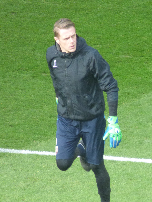Ein Mann in einer schwarzen Jacke und blauen Shorts läuft auf einem Fußballfeld und trägt Fußballhandschuhe.