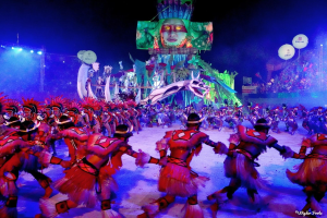 Eine Gruppe von Menschen in bunten Kostümen tanzt auf einer Bühne vor einem sitzenden Publikum, mit Lichtern, Pfählen, Ballons und einem Hintergrundbild, unter einem sichtbaren Himmel, bei der Rio-Karneval 2014.