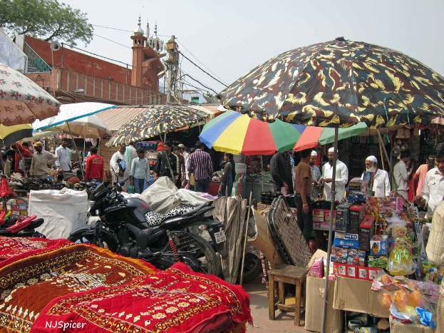Eine belebte Szene mit verschiedenen Gegenständen, darunter Schirme, Fahrzeuge, Matten, Kisten, einen Hocker, Menschen, ein Mikrofon, eine Stromstange, Kabel und eine Moschee im Hintergrund.