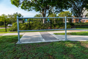Ein Metallzaun in einer grasbewachsenen Fläche mit Pflanzen, Bäumen und einem Auto auf der Straße, mit Gebäuden und einem klaren blauen Himmel im Hintergrund.