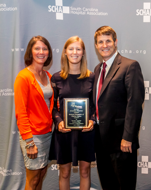 Drei Personen, zwei Frauen und ein Mann, die zusammen stehen, wobei eine Frau einen Preis hält, vor einem grauen Hintergrund.