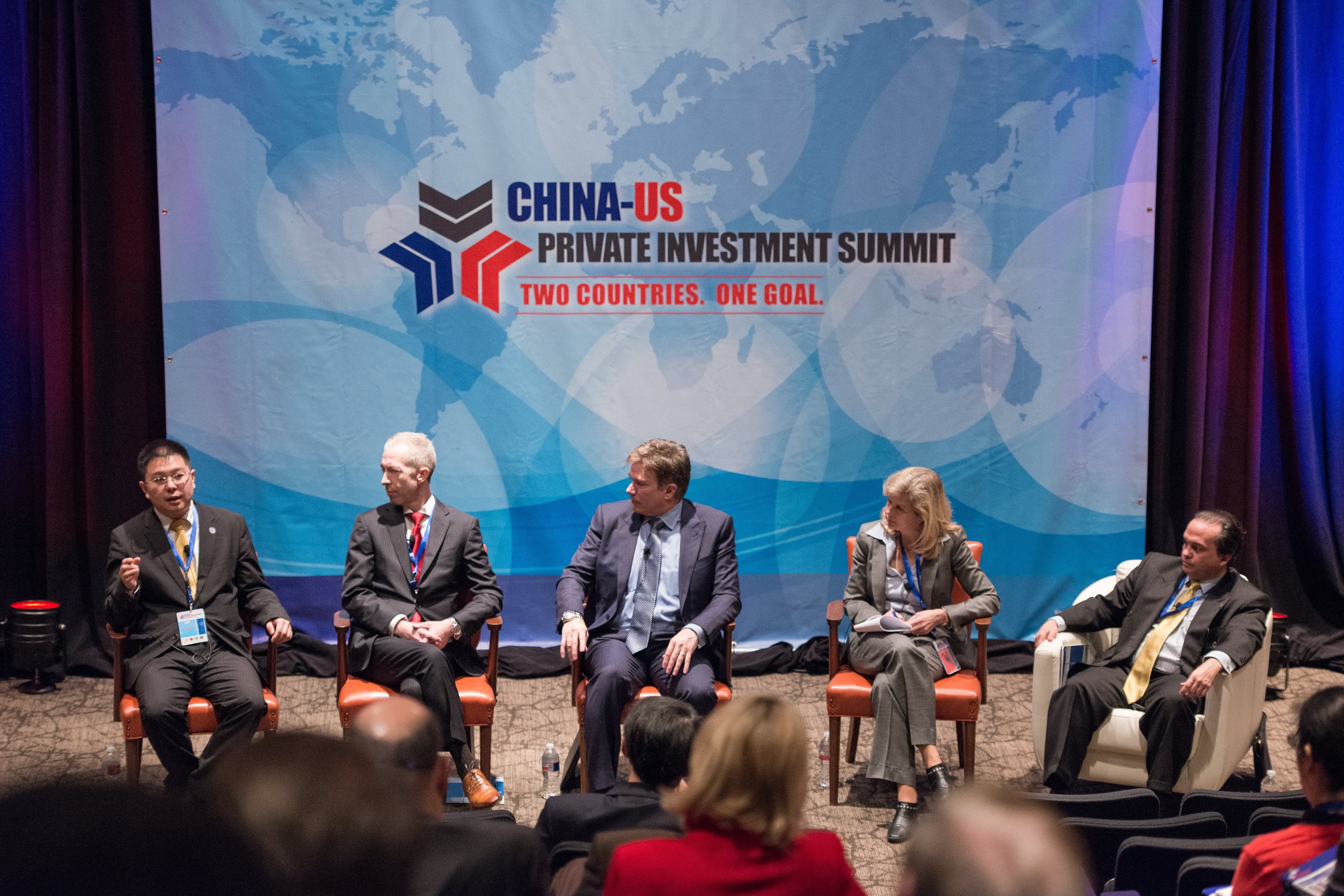 Gruppe von Menschen, die auf Stühlen sitzen und einem Bildschirm mit Text und einer Karte gegenüberliegen, mit Vorhängen im Hintergrund, was auf einen China-US-Privatinvestment-Gipfel hindeutet.