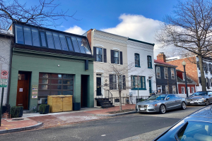 Eine Straße in einem Stadtteil von Philadelphia, gesäumt von parkenden Autos, Gebäuden, Bäumen, Laternen, Schildern und Holzboxen unter einem bewölkten Himmel.