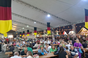 Eine große Gruppe von Menschen sitzt an Tischen unter einem Zelt auf dem Oktoberfest in München, Deutschland, mit einigen Stehenden und Taschen auf den Tischen, umgeben von Zelten, Lichtern und Fahnen.