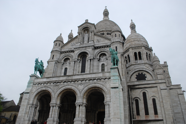 Ein Denkmal steht in der Mitte des Bildes, mit Häusern und einem Baum in der unteren linken Ecke.