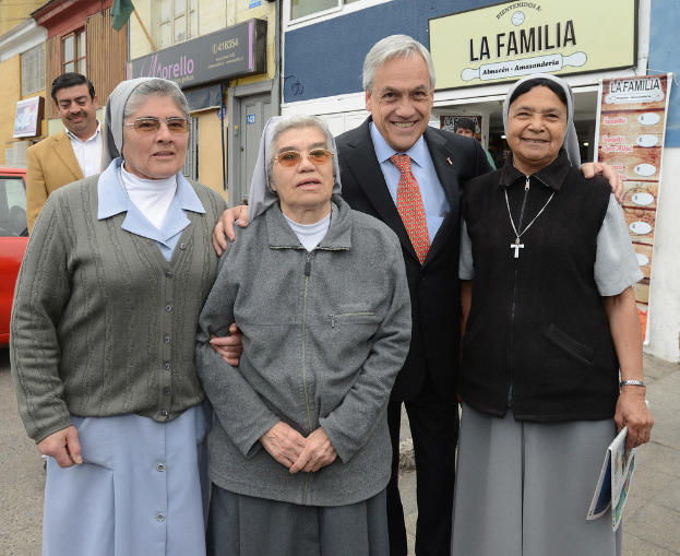 Eine Gruppe von Menschen, die lachend vor einem Gebäude mit einer Wand, einer T√ľr, Fahnen, einem Auto und einer weiteren Person im Hintergrund steht.