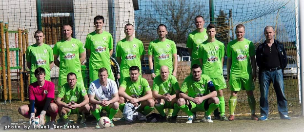 Eine Gruppe von Männern in grünen T-Shirts steht auf einem Fußballfeld, einige hocken, mit einem Ball vor ihnen und einem Torpfosten rechts; der Hintergrund enthält Bäume, Gebäude und Fahrzeuge.