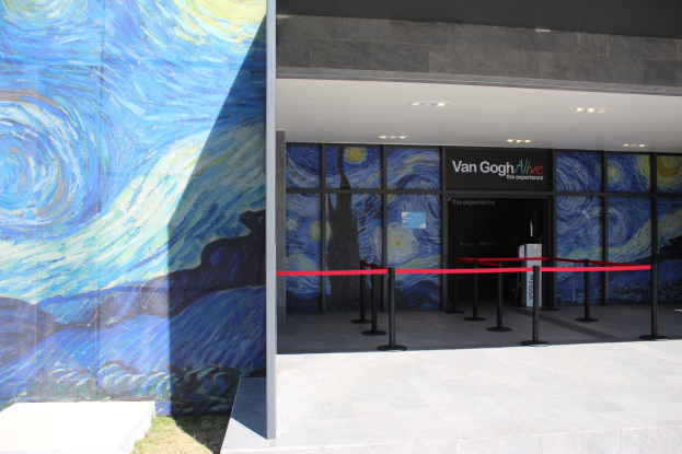 Der Eingang eines Gebäudes zeigt ein großes Wandgemälde, ein Schild mit der Aufschrift "Van Gogh Alive", Absperrbaken mit Bändern, Gras auf dem Boden und Lampen an der Decke.