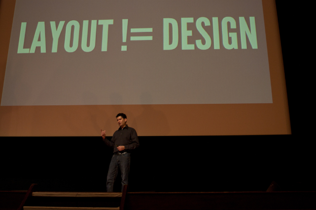 Eine Person steht auf einer Bühne und spricht, mit einem Bildschirm im Hintergrund, auf dem Text angezeigt wird.