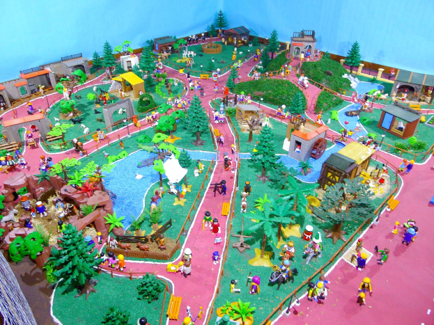 Ein Miniaturzoo-Modell bevölkert mit Menschen, umgeben von Bäumen, Häusern, Pflanzen, Gras, einem Zaun und anderen Gegenständen, mit einer Wand im Hintergrund und Text unten.