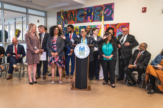 Gruppe von Menschen vor einem Podium mit einem Mikrofon und einer Tafel mit dem Text "Sancho City Councilman Joins City Council", mit sitzenden und stehenden Zuschauern, Flaggen, Feuerlöscher und Deckenleuchten im Hintergrund.