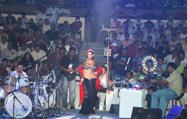 Eine Person spielt ein Musikinstrument auf der Bühne, ein Mann in einem schwarzen Hemd hält ein Mikrofon, eine Frau in einem schwarzen und roten Kleid steht neben ihm, eine Person sitzt in der Ecke und hält eine Kamera, das Publikum sitzt auf Stühlen, ein Tisch mit Gläsern und Flaschen steht vor dem Publikum und ein Lautsprecher ist unten im Bild.