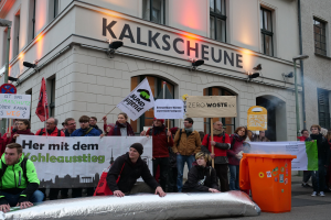 Eine Gruppe von Menschen demonstriert vor einem Gebäude in Deutschland, hält Schilder und Plakate hoch und hat zwei Personen im Vordergrund und einen Müllcontainer auf der rechten Seite.